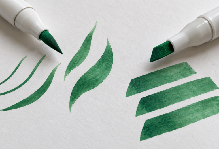 Primer plano de dos rotuladores blancos con puntas pincel y biselada junto a trazos verdes finos, anchos y curvos.